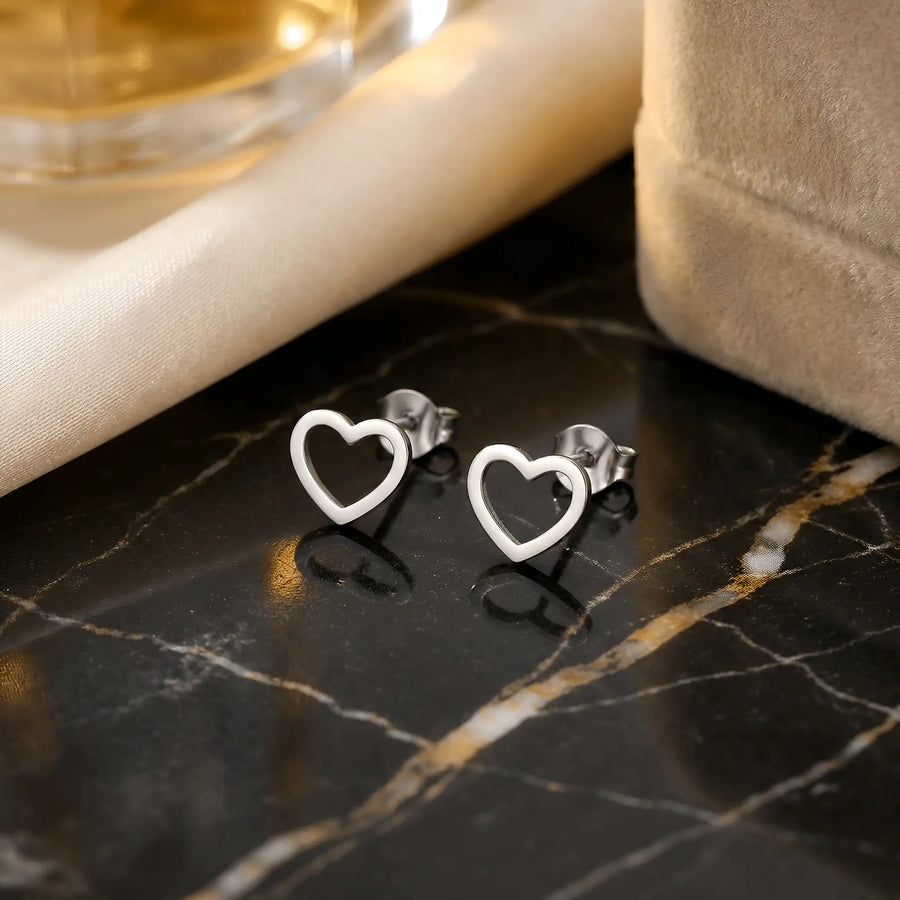 Cercei argint 925 rodiat stud cu inimă outline 9x8mm — pereche pe placă de marmură neagră lăcuită cu pernuță bej, cadrul subțire al inimii vizibil clar, finisaj oglindă alb-platinat, prindere fluturaș push-back