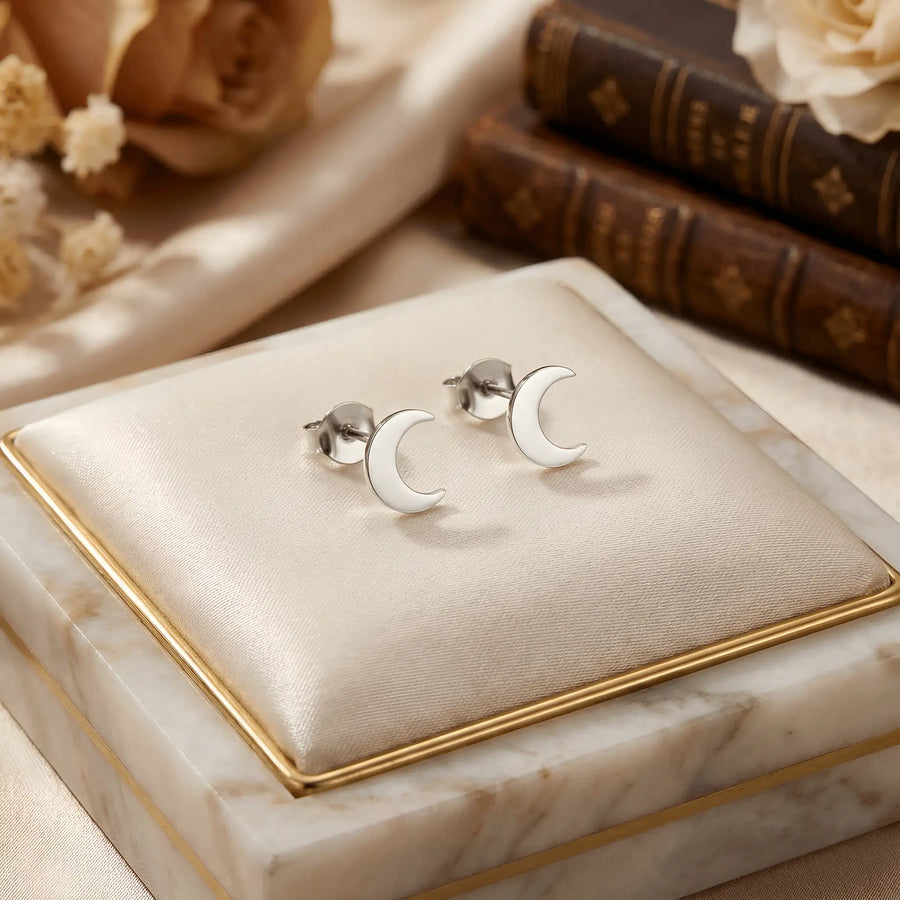 Cercei argint 925 rodiat stud cu semilună plină 9x7mm — pereche pe pernuță crem cu marmură albă cu margine aurie, cărți vechi cu cotor maro și flori uscate trandafiri pudrați în fundal, finisaj oglindă alb-platinat