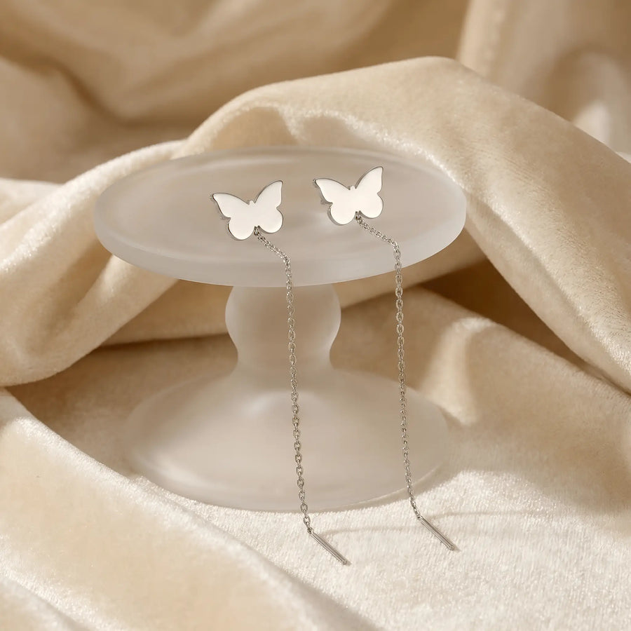 Cercei argint 925 rodiat threader cu fluture plin stilizat 7,5mm — pereche pe suport rotund de sticlă mată cu drape velvet crem-bej, aripi pline simetrice, corp central subțire, lanț 6cm cu tijă-bară, finisaj oglindă alb-platinat