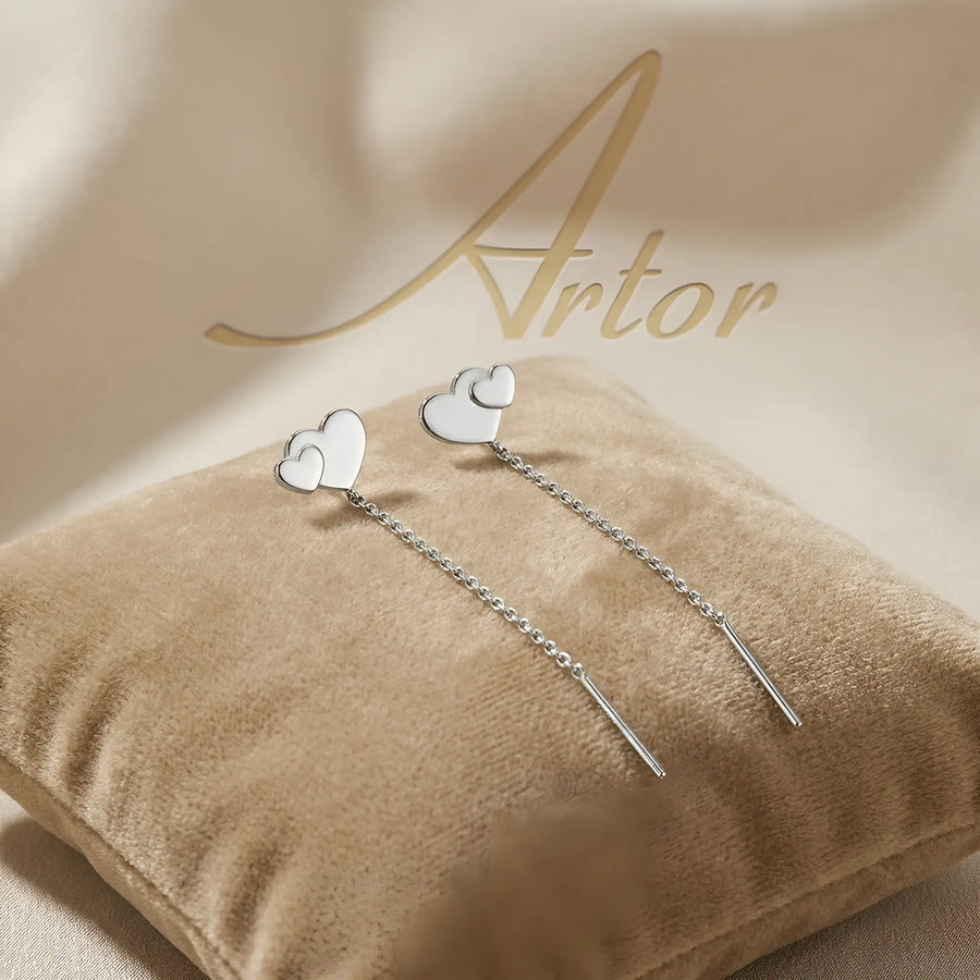 Cercei argint 925 rodiat threader cu inimă dublă suprapusă — pereche pe pernă velvet bej cu logo Artor pe fundal satin, două inimi pline atinse, lanț subțire 6cm cu tijă-bară, finisaj oglindă alb-platinat