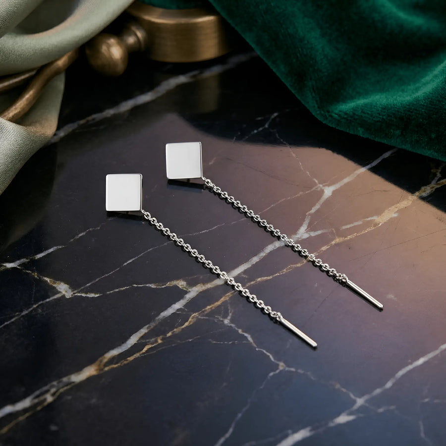 Cercei argint 925 rodiat threader cu pătrat plin geometric 9x9mm — pereche pe placă marmură neagră cu vinete aurii, draperie velvet verde smarald în fundal, accente metal alămit, lanț subțire 6cm cu tijă-bară, finisaj oglindă alb-platinat