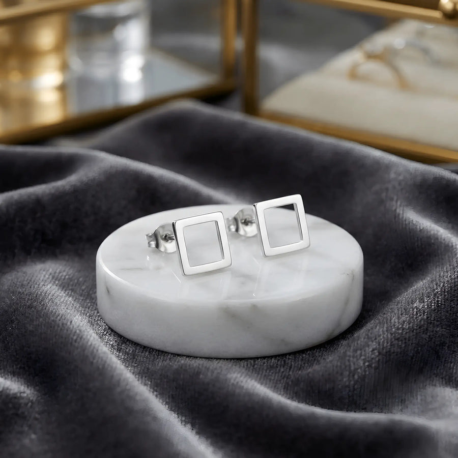 Cercei argint 925 rodiat stud cu pătrat outline 6x6mm — pereche pe placă rotundă de marmură albă pe pânză gri velvet, cutii de bijuterii cu detalii aurii pe fundal, finisaj oglindă alb-platinat, prindere fluturaș push-back