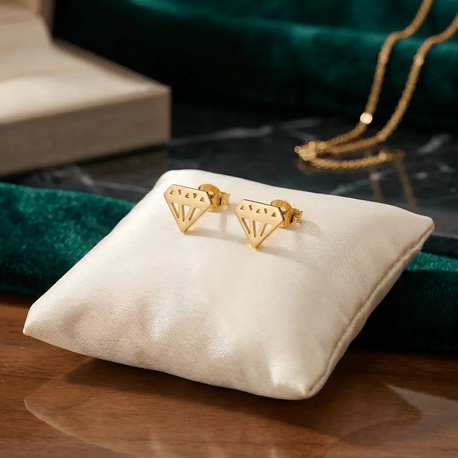 Cercei aur 14K stud diamant stilizat geometric — vedere produs cu siluetă brilliant decupată cu fațete pe pernă albă, fundal verde catifea