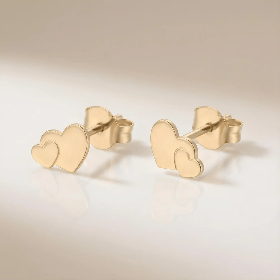 Cercei aur 14K stud cu inimă dublă suprapusă — pereche pe fundal crem, două inimi pline una mai mică peste una mai mare, finisaj oglindă, prindere fluturaș push-back, dimensiune 11x7.5mm