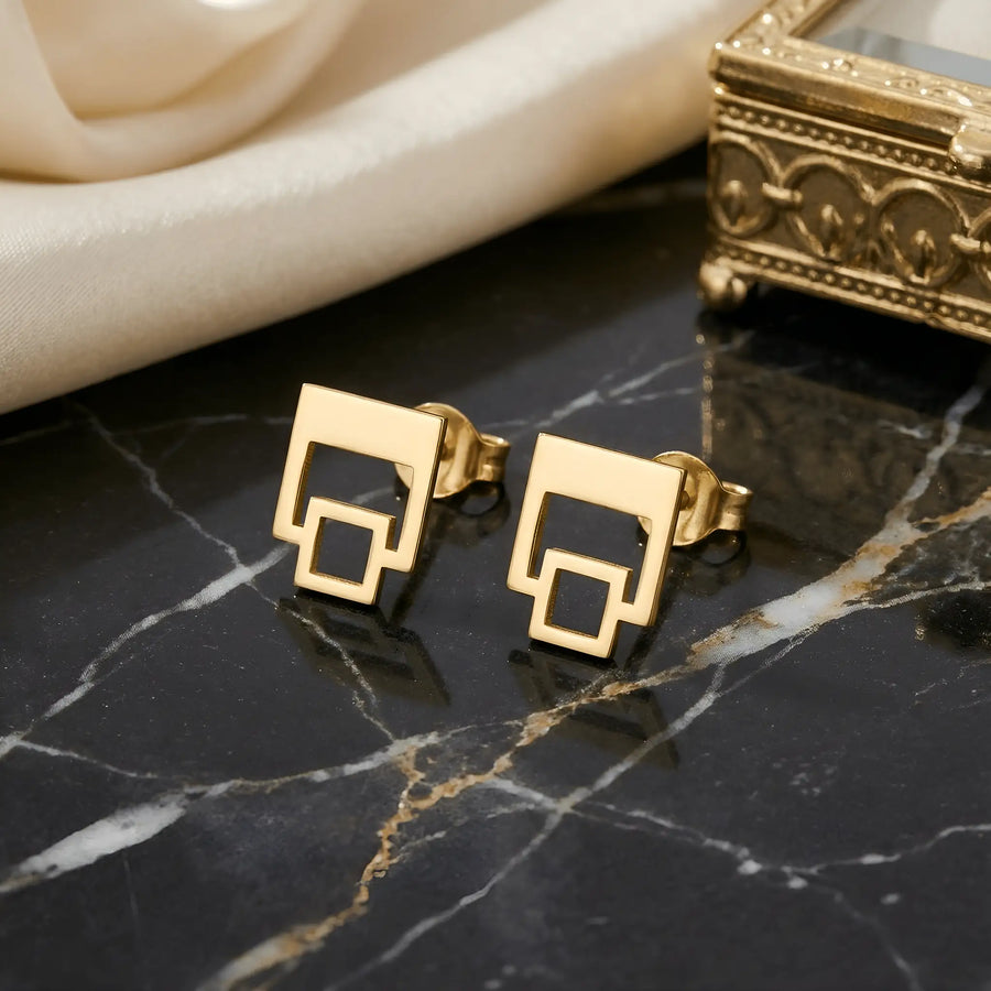 Cercei aur 14K stud cu două pătrate outline suprapuse 9,3mm — pereche pe placă marmură neagră cu vinete aurii, draperie satin crem și cutie ornamentală aurie pe fundal, pătrat mare sus și pătrat mic atașat jos, finisaj oglindă, prindere fluturaș clasic