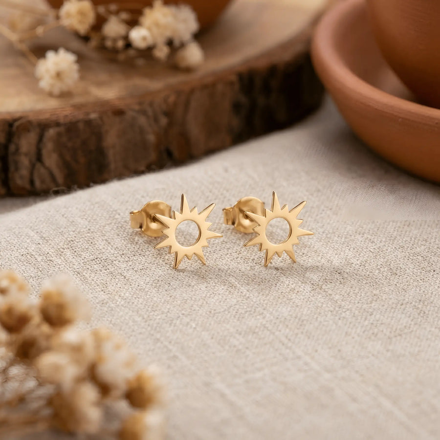 Cercei aur 14K stud cu soare stilizat 10x10mm — pereche pe pânză crem cu boluri de teracota și flori uscate gypsophila, soare cu cerc central decupat și 8 raze alternate, finisaj oglindă