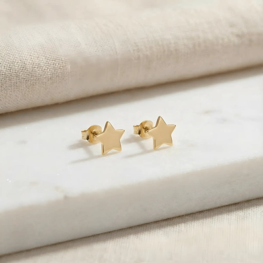 Cercei aur 14K stud cu stea mică plină 6x6mm — pereche pe placă de marmură albă cu pânză crem fundal, două steluțe simetrice cu 5 colțuri, finisaj oglindă, prindere fluturaș push-back vizibilă