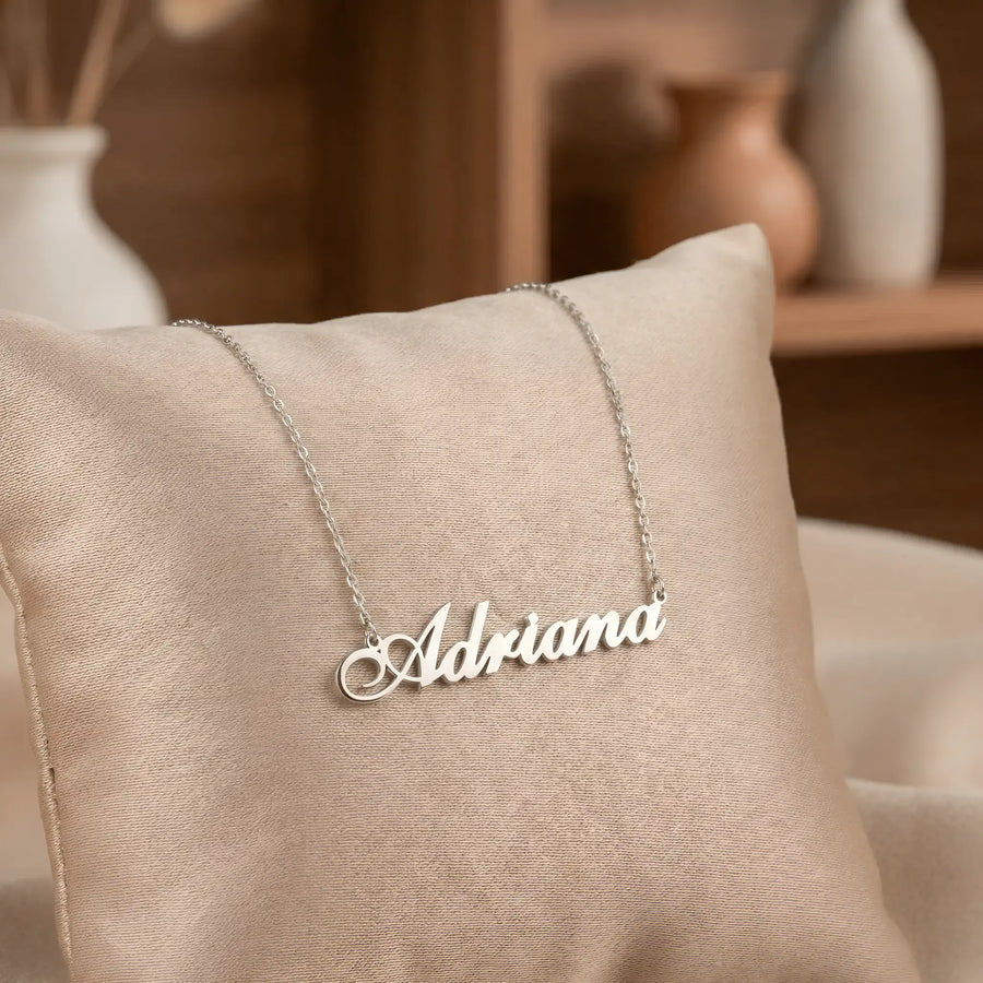 Lănțișor argint 925 rodiat cu numele Adriana, pandantiv decupat 42x10 mm font cursiv inițiala A cu buclă jos-stânga, lanț anchor 46 cm pe pernuță textilă bej cu vază ceramică pe raft de lemn