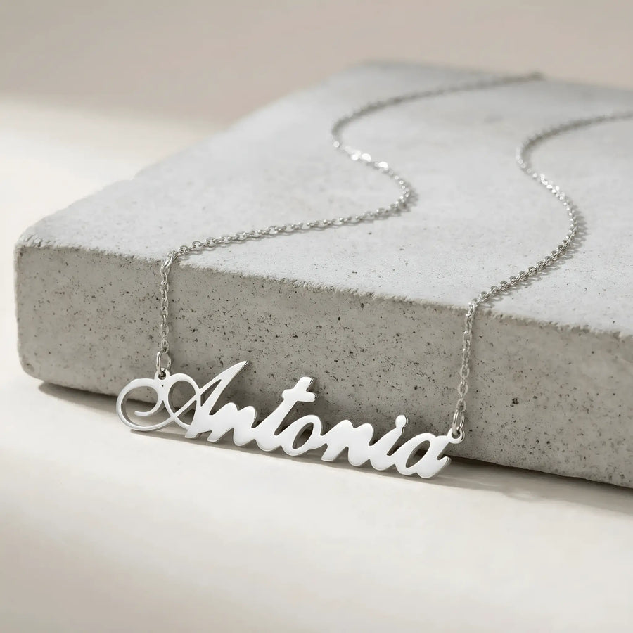 Lănțișor argint 925 rodiat personalizat cu numele Antonia decupat în scriere cursivă elegantă cu coadă caligrafică pe placă de beton gri, lanț subțire alb-platinat