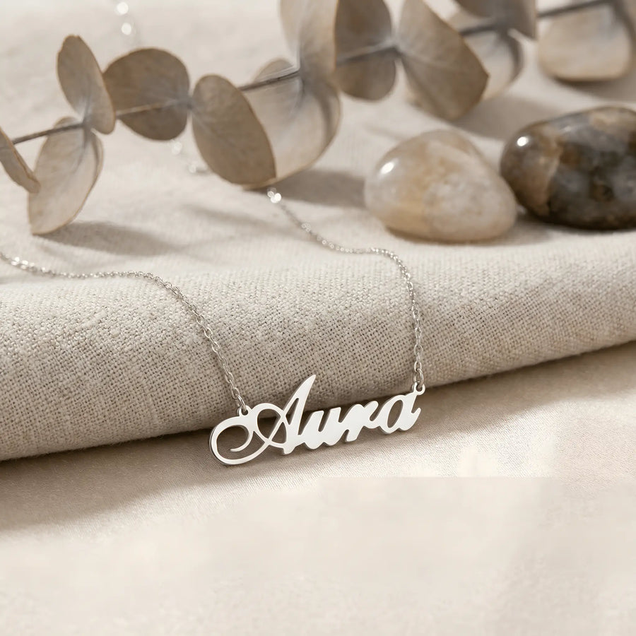 Lănțișor argint 925 rodiat personalizat cu numele Aura decupat în scriere cursivă elegantă cu A larg și coadă caligrafică pe textil bej cu eucalipt și pietre, lanț subțire alb-platinat