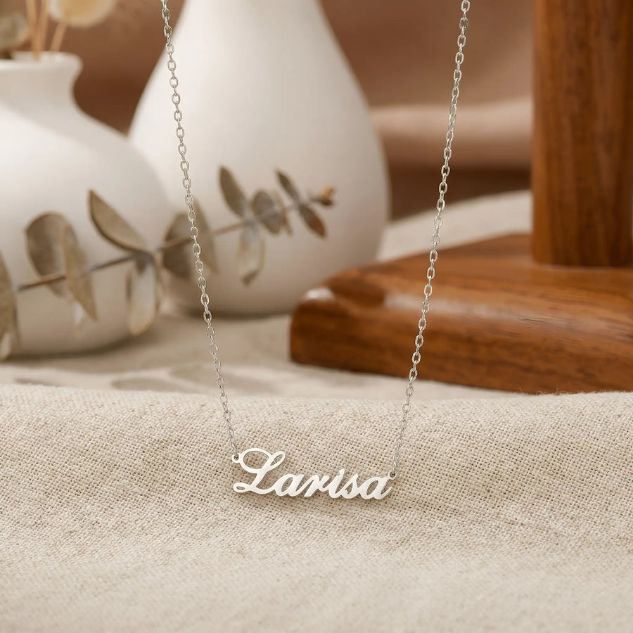 Lănțișor argint 925 rodiat personalizat cu numele Larisa decupat în scriere cursivă elegantă cu L larg pe textil bej cu vase ceramice albe și eucalipt, lanț subțire alb-platinat