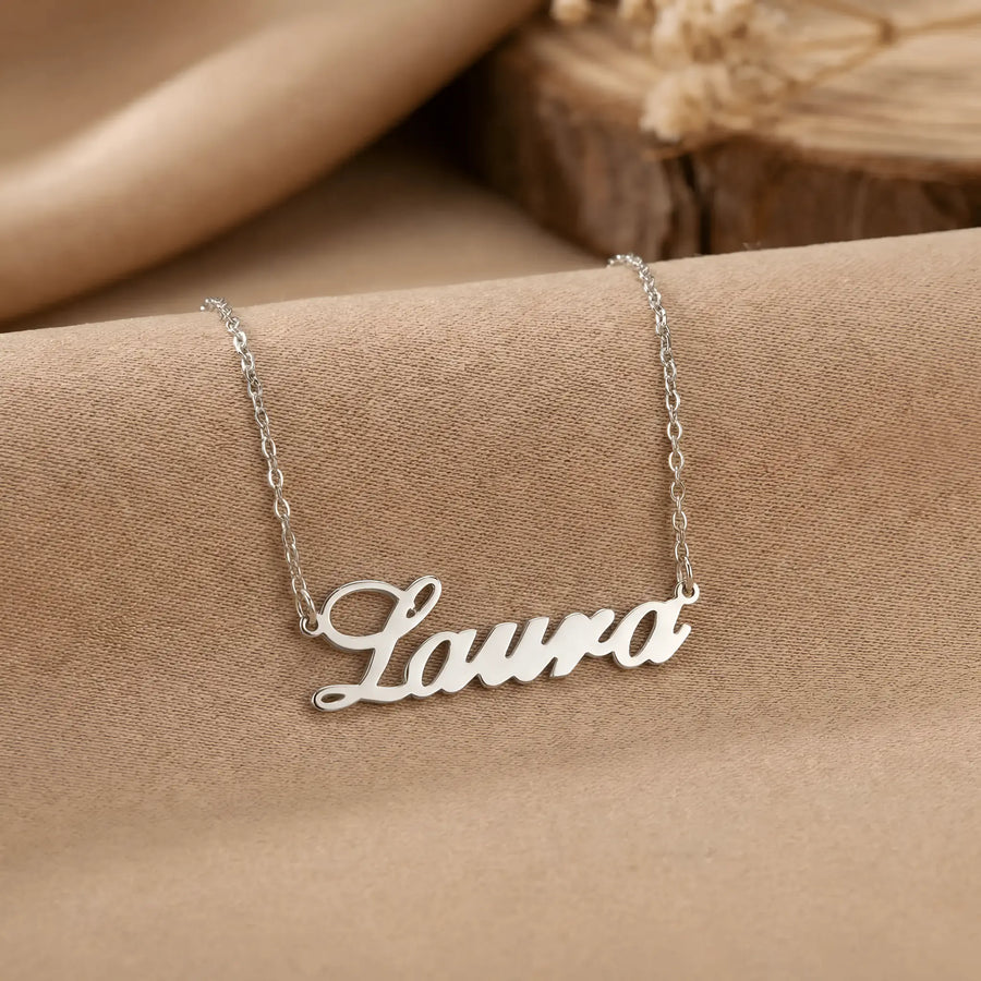 Lănțișor argint 925 rodiat personalizat cu numele Laura decupat în scriere cursivă elegantă cu L larg pe textil bej cu suport lemn rotund, lanț subțire alb-platinat