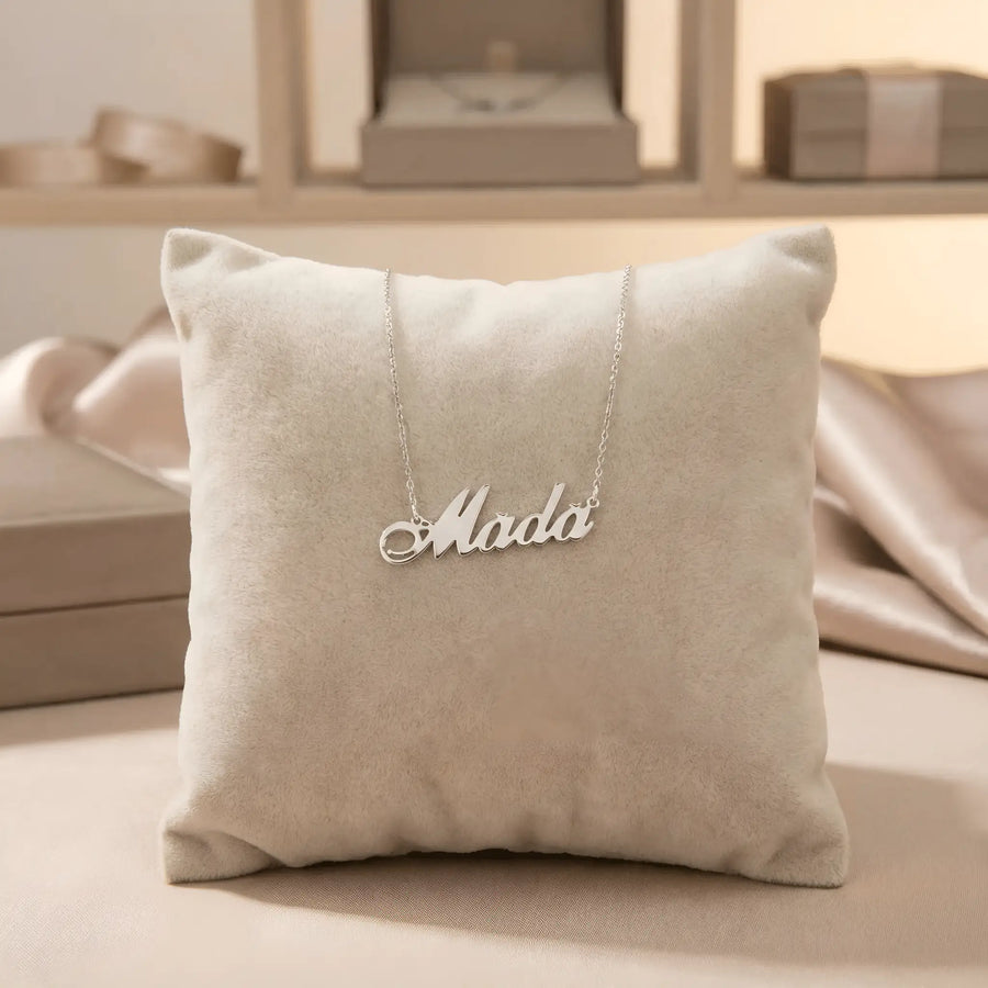 Lănțișor argint 925 rodiat personalizat cu numele Mada decupat în scriere cursivă elegantă cu M cu coadă caligrafică pe pernuță bej display velvet, lanț subțire alb-platinat