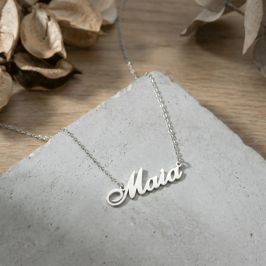 Lănțișor argint 925 rodiat personalizat cu numele Maia decupat în scriere cursivă elegantă cu M cu coadă caligrafică pe stâncă gri pe lemn cu frunze uscate, lanț subțire alb-platinat