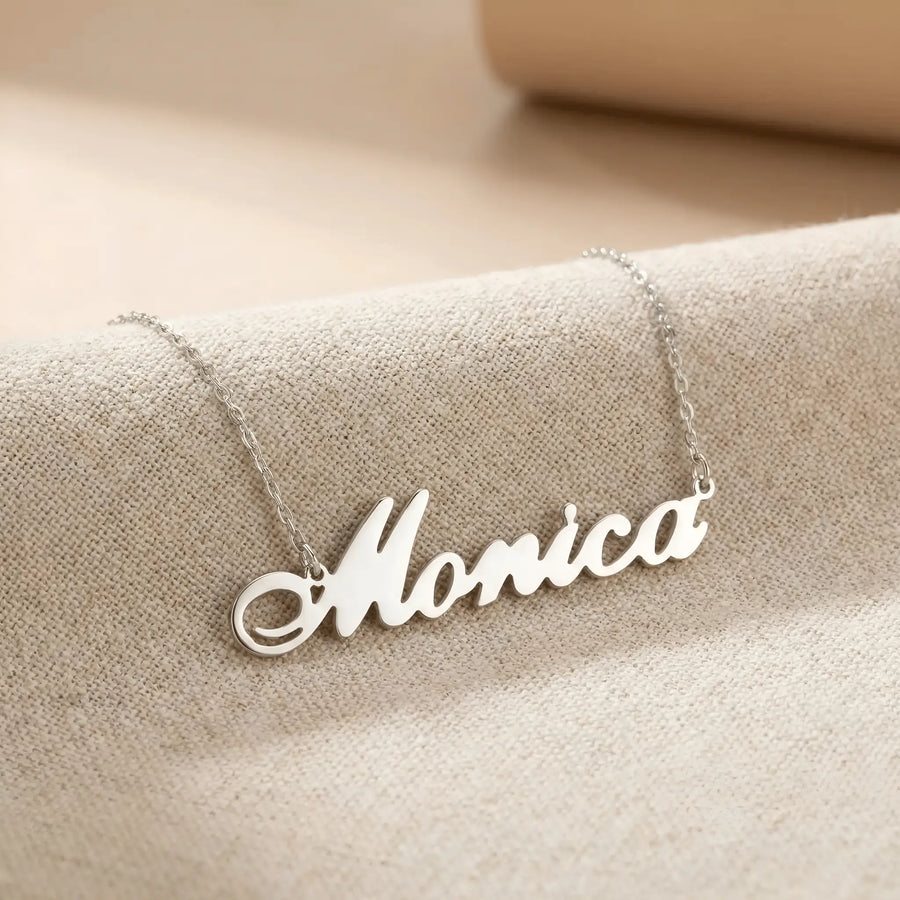 Lănțișor argint 925 rodiat personalizat cu numele Monica decupat în scriere cursivă elegantă cu M larg pe textil bej, lanț subțire alb-platinat