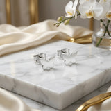 Cercei argint 925 rodiat stud cu fluture plin 10x6.5mm — pereche pe placă de marmură albă cu pânză mătăsoasă crem și flori de orhidee albă pe fundal, fluturași simetrici cu silueta plină, finisaj oglindă alb-platinat