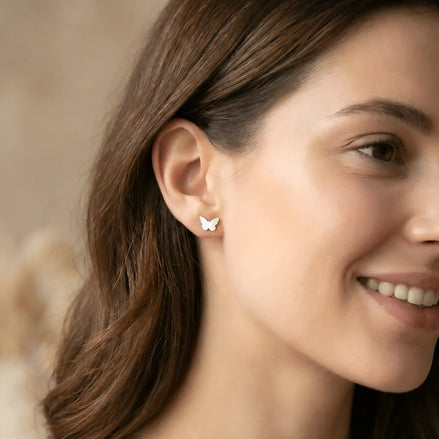 Cercei argint 925 rodiat stud fluture — purtată pe lobul urechii unei tinere brunete zâmbitoare cu păr lung castaniu, fluture mic vizibil pe ureche, ten luminos, fundal cald natural