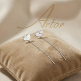 Cercei argint 925 rodiat threader cu inimă dublă suprapusă — pereche pe pernă velvet bej cu logo Artor pe fundal satin, două inimi pline atinse, lanț subțire 6cm cu tijă-bară, finisaj oglindă alb-platinat