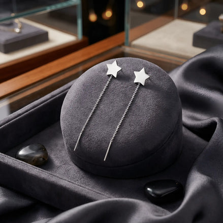 Cercei argint 925 rodiat threader cu stea mare plină 8,5mm — pereche pe pernă rotundă velvet gri-grafit cu satin gri închis pe fundal, pietre semi-prețioase obsidian negre, vitrina cu bijuterii pe lateral, lanț 6cm cu tijă-bară, finisaj oglindă alb-platinat