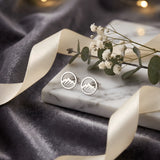 Cercei argint 925 rodiat stud cu munte în cerc outline 8,6mm — pereche pe placă marmură albă pe drape velvet gri-grafit cu satin auriu și lumini bokeh aurii pe fundal, ramuri eucalipt și flori gypsophila în spate, finisaj oglindă alb-platinat, prindere fluturaș clasic