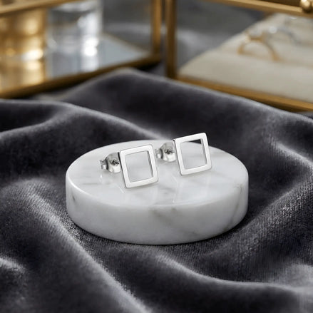 Cercei argint 925 rodiat stud cu pătrat outline 6x6mm — pereche pe placă rotundă de marmură albă pe pânză gri velvet, cutii de bijuterii cu detalii aurii pe fundal, finisaj oglindă alb-platinat, prindere fluturaș push-back