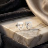 Cercei argint 925 rodiat stud cu dublu pătrat outline 9,3mm — pereche pe placă marmură albă cu vinete maro pe drape velvet gri-grafit cu satin auriu pe fundal, pătrat mare sus și pătrat mic atașat în jos cu interior gol, finisaj oglindă alb-platinat, prindere fluturaș clasic