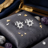 Cercei argint 925 rodiat stud cu soare stilizat 10x10mm — pereche pe pernuță gri-velvet cu pietre de cristal de stâncă, ametist și fragmente aurii decorative, cutie de bijuterii cu detalii aurii pe fundal, finisaj oglindă alb-platinat