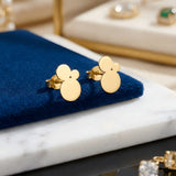 Cercei aur 14K stud cu trio de cercuri pline suprapuse 11mm — pereche pe pernă velvet albastru-navy cu placă marmură albă, vitrina cu bijuterii cu pietre verzi smarald pe fundal, siluetă verticală cu trei cercuri descrescătoare, finisaj oglindă, prindere fluturaș clasic