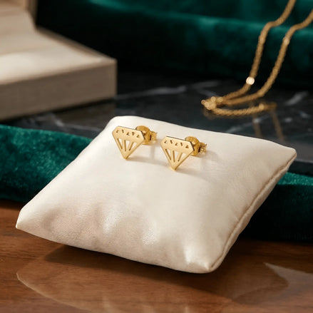 Cercei aur 14K stud diamant stilizat geometric — vedere produs cu siluetă brilliant decupată cu fațete pe pernă albă, fundal verde catifea