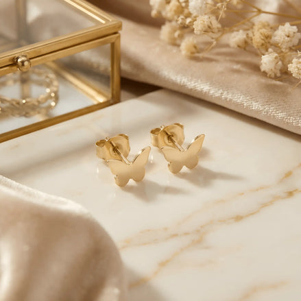 Cercei aur 14K stud cu fluture plin — pereche pe fundal de marmură crem cu cutie de bijuterii din sticlă, fluturi mici lustruiți oglindă, prindere fluturaș push-back, ramuri de gypsophila albă