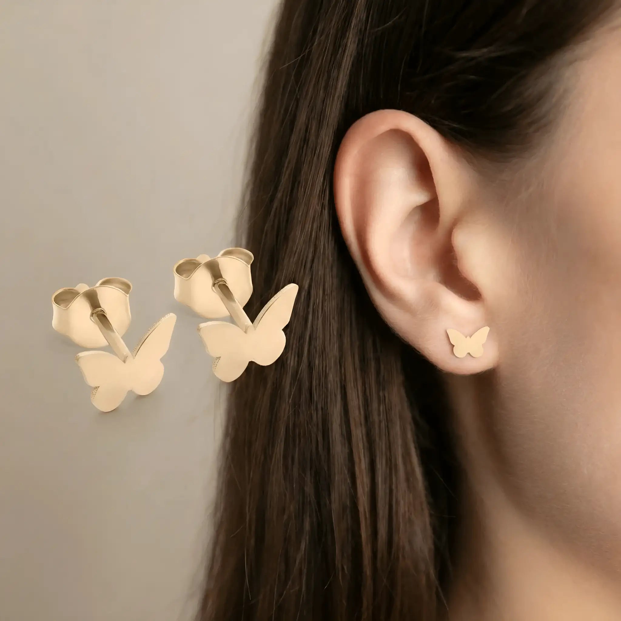 Cercei aur 14K stud fluture — purtată pe lobul urechii unei tinere brunete, fluturele mic vizibil clar pe ureche, păr lung castaniu cădere natural