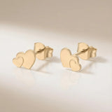 Cercei aur 14K stud cu inimă dublă suprapusă — pereche pe fundal crem, două inimi pline una mai mică peste una mai mare, finisaj oglindă, prindere fluturaș push-back, dimensiune 11x7.5mm
