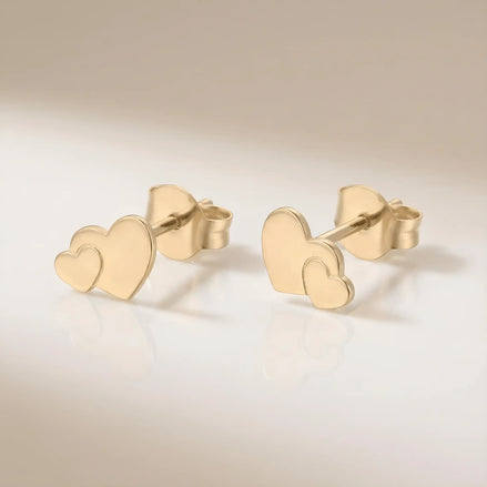 Cercei aur 14K stud cu inimă dublă suprapusă — pereche pe fundal crem, două inimi pline una mai mică peste una mai mare, finisaj oglindă, prindere fluturaș push-back, dimensiune 11x7.5mm