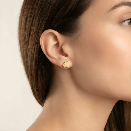 Cercei aur 14K stud inimă dublă purtați pe ureche — model damă păr brunet, bijuterie compactă pe lob cu cele două inimi suprapuse vizibile lateral