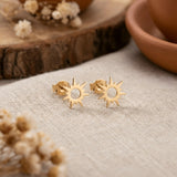 Cercei aur 14K stud cu soare stilizat 10x10mm — pereche pe pânză crem cu boluri de teracota și flori uscate gypsophila, soare cu cerc central decupat și 8 raze alternate, finisaj oglindă
