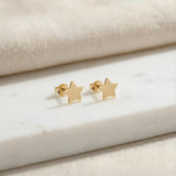 Cercei aur 14K stud cu stea mică plină 6x6mm — pereche pe placă de marmură albă cu pânză crem fundal, două steluțe simetrice cu 5 colțuri, finisaj oglindă, prindere fluturaș push-back vizibilă