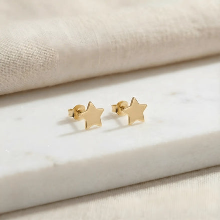 Cercei aur 14K stud cu stea mică plină 6x6mm — pereche pe placă de marmură albă cu pânză crem fundal, două steluțe simetrice cu 5 colțuri, finisaj oglindă, prindere fluturaș push-back vizibilă