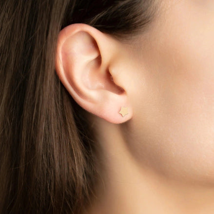 Cercei aur 14K stud stea mică — purtată pe lobul urechii unei tinere brunete, steluța micuță abia vizibilă pe ureche, păr lung castaniu cădere natural