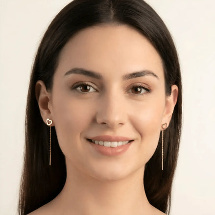 Cercei aur 14K threader inimă contur purtați pe ureche — model dama păr brunet zâmbind, conturul subțire al inimii deasupra lobului, lănțișor pe spate