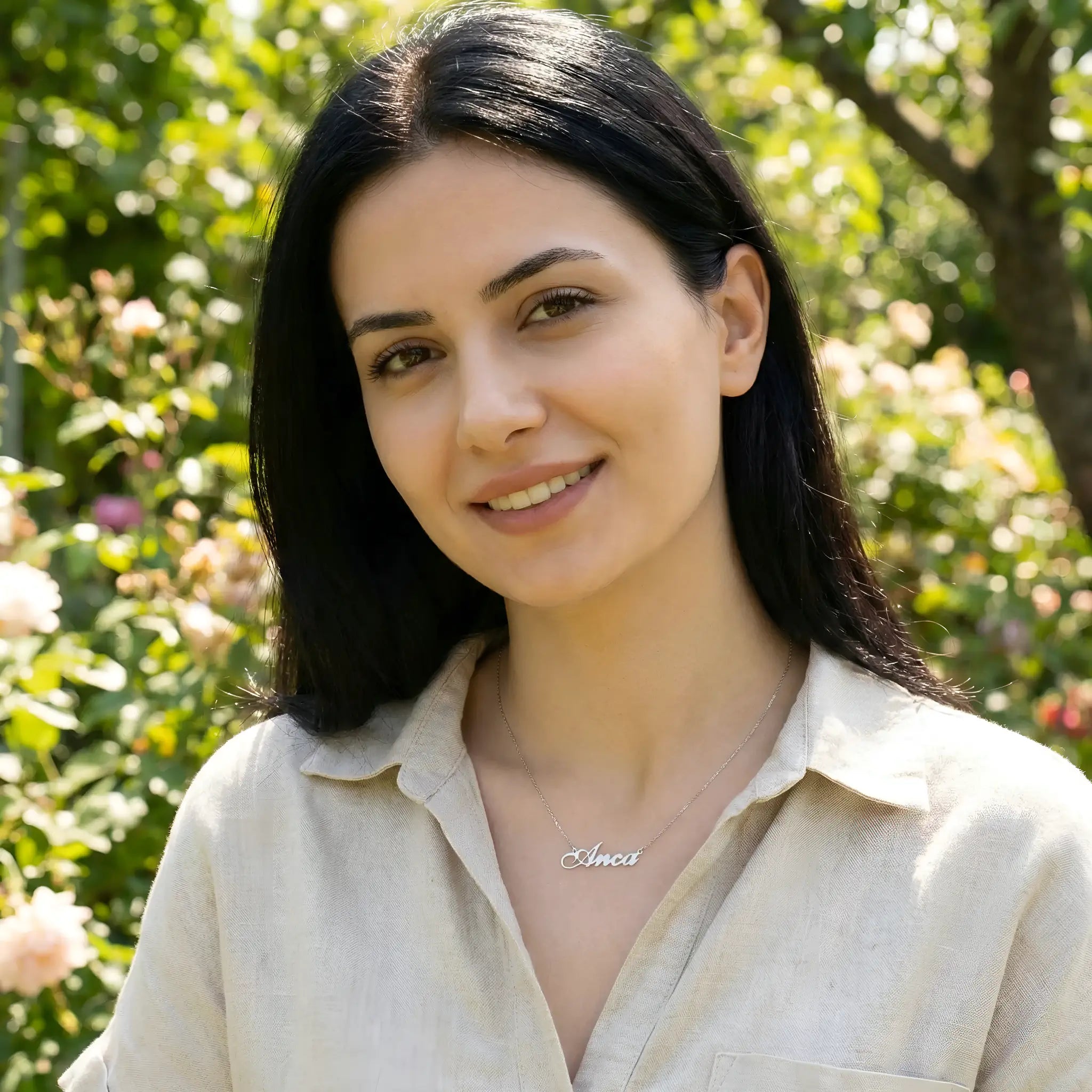 Damă tânără purtând lănțișor argint 925 rodiat cu numele Anca script, peste cămașă bej de in, lumină naturală exterior cu vegetație de vară și flori