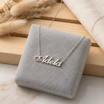 Lănțișor argint 925 rodiat cu numele Adela, pandantiv decupat 31x10 mm font cursiv inițiala A cu buclă jos-stânga, lanț anchor 46 cm pe cutie velvet gri cu fundal de iarbă pampas uscată