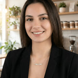 Damă cu păr lung negru purtând lănțișor argint 925 rodiat Adelina pe stern peste sacou negru desfăcut, cafenea cu plante și râșniță cafea