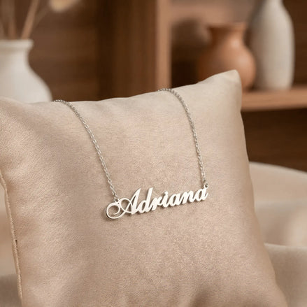 Lănțișor argint 925 rodiat cu numele Adriana, pandantiv decupat 42x10 mm font cursiv inițiala A cu buclă jos-stânga, lanț anchor 46 cm pe pernuță textilă bej cu vază ceramică pe raft de lemn