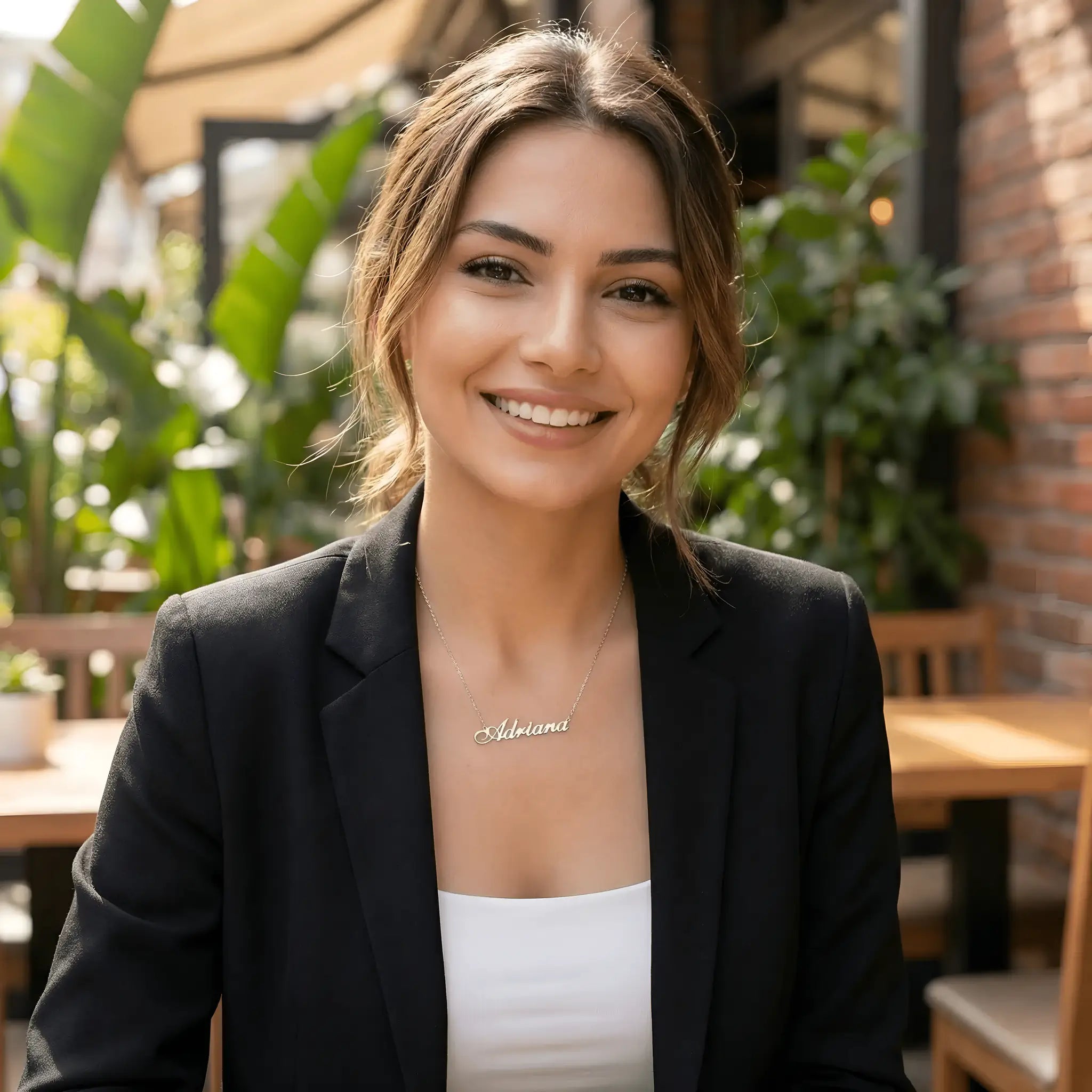 Damă cu păr lung castaniu prins lejer purtând lănțișor argint 925 rodiat Adriana deasupra claviculei peste sacou negru și top alb, terasă cu plante tropicale și zid de cărămidă