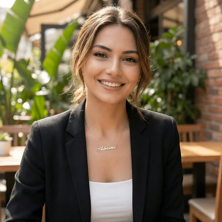 Damă cu păr lung castaniu prins lejer purtând lănțișor argint 925 rodiat Adriana deasupra claviculei peste sacou negru și top alb, terasă cu plante tropicale și zid de cărămidă
