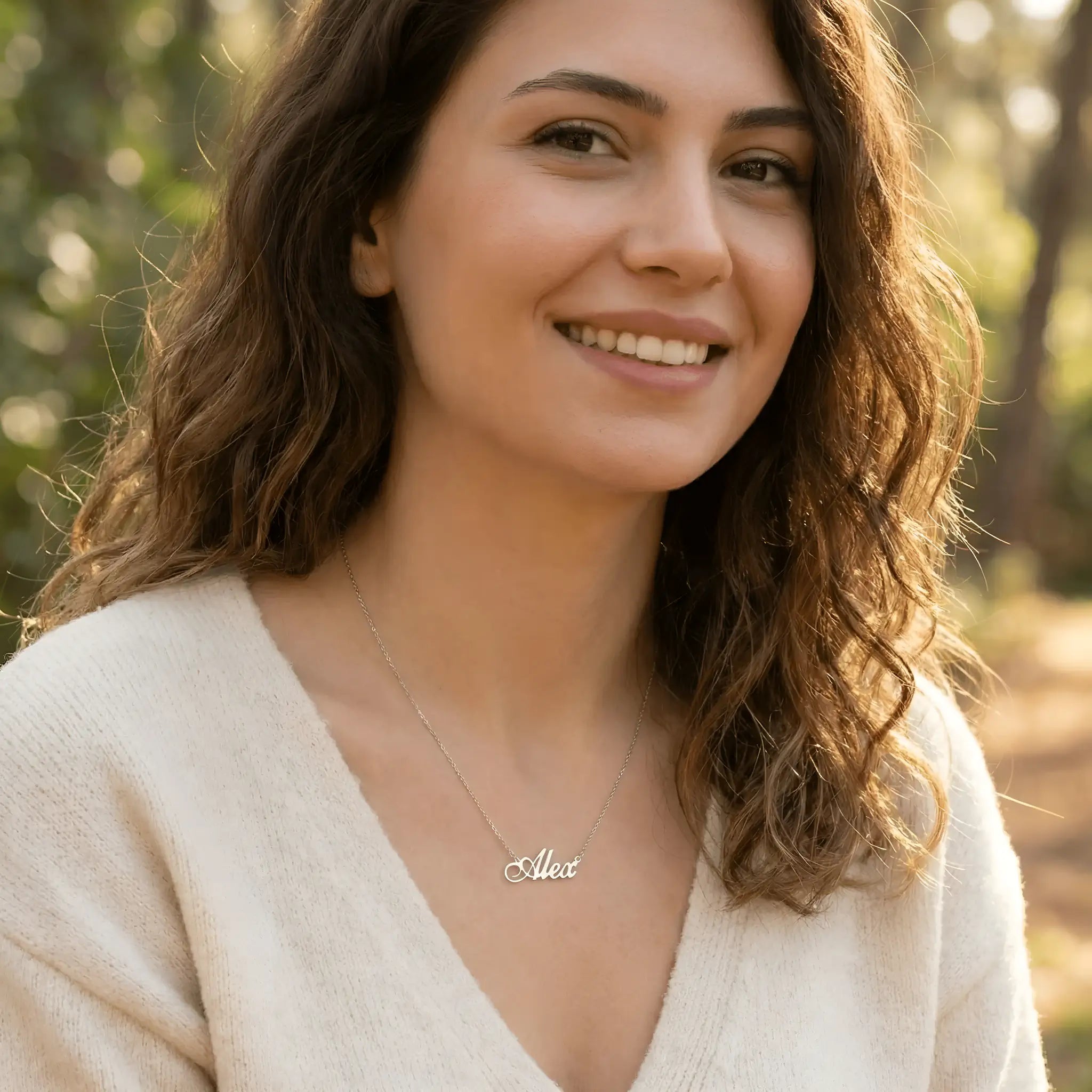 Damă tânără purtând lănțișor argint 925 rodiat cu numele Alex script, peste pulover crem fin cu decolteu V, lumină naturală exterior cu vegetație