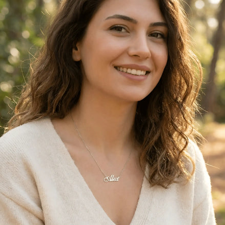 Damă tânără purtând lănțișor argint 925 rodiat cu numele Alex script, peste pulover crem fin cu decolteu V, lumină naturală exterior cu vegetație