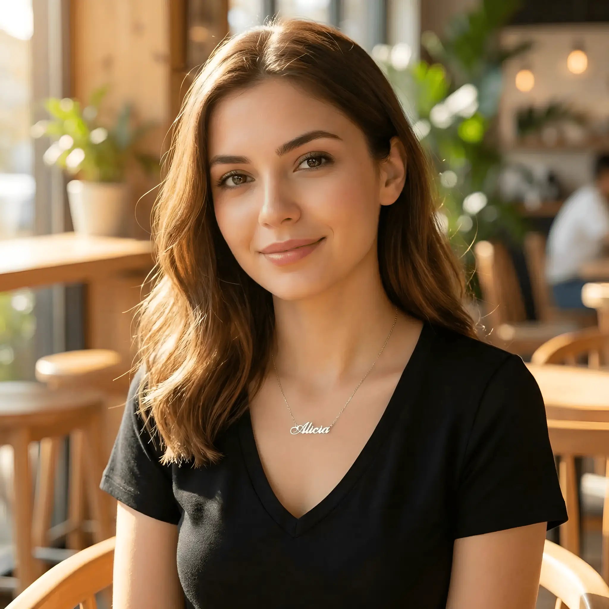Damă tânără purtând lănțișor argint 925 rodiat cu numele Alicia script, peste t-shirt negru cu decolteu V, cafenea cu lumină caldă și mobilier de lemn