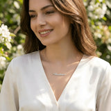 Damă cu păr lung castaniu-roșcat purtând lănțișor argint 925 rodiat Anastasia deasupra claviculei peste bluză satinată ivory cu decolteu V, grădină cu flori albe lumină naturală caldă