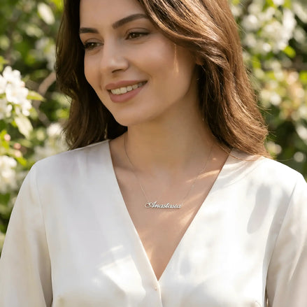Damă cu păr lung castaniu-roșcat purtând lănțișor argint 925 rodiat Anastasia deasupra claviculei peste bluză satinată ivory cu decolteu V, grădină cu flori albe lumină naturală caldă
